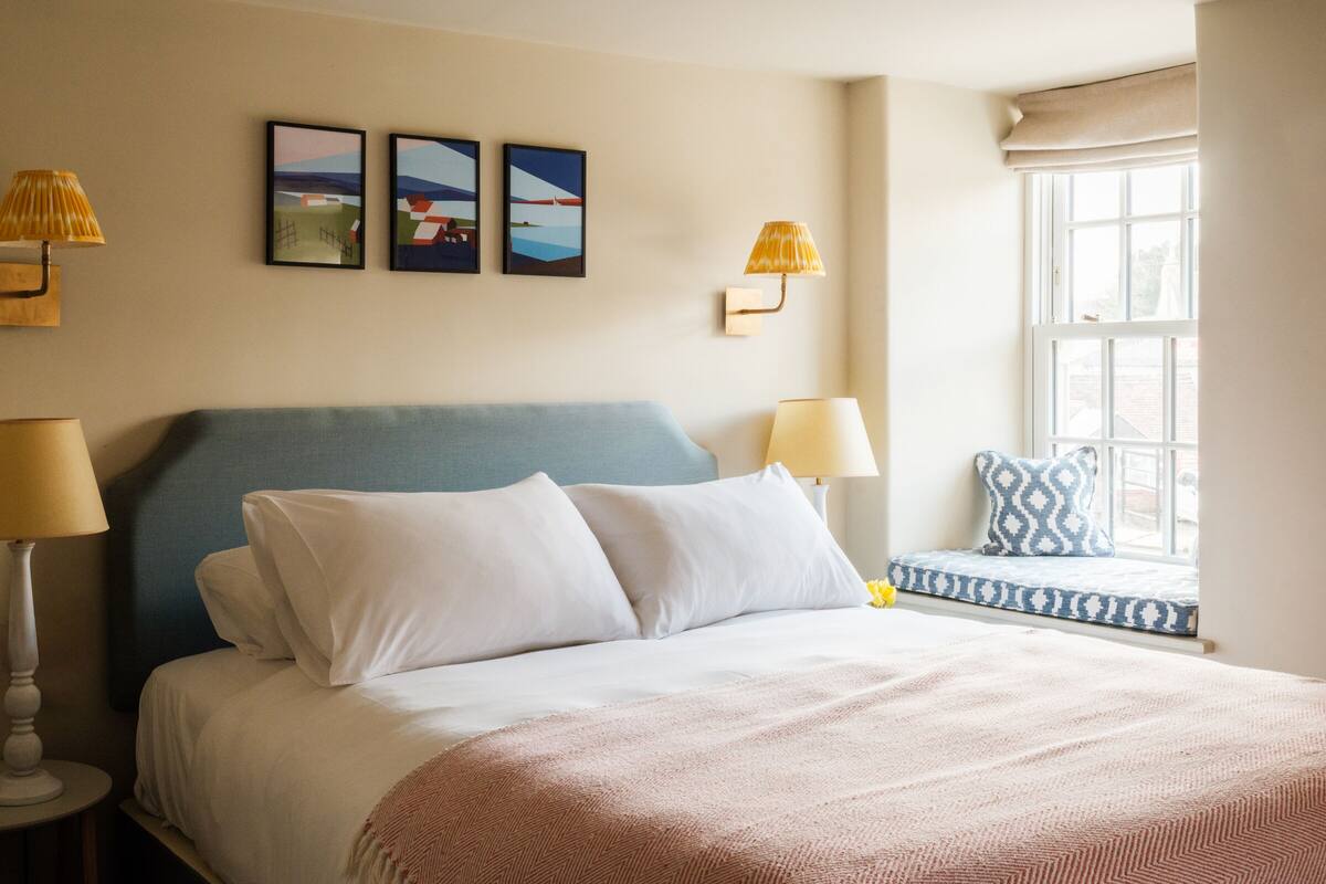 A bedroom with a bed, two wall-mounted lamps, a window with a view, and three framed pictures on the wall.