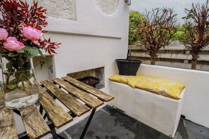A balcony with a wooden table, a bench with a yellow cushion, a wicker basket, and a vase with pink flowers. - Penvar (Truro)