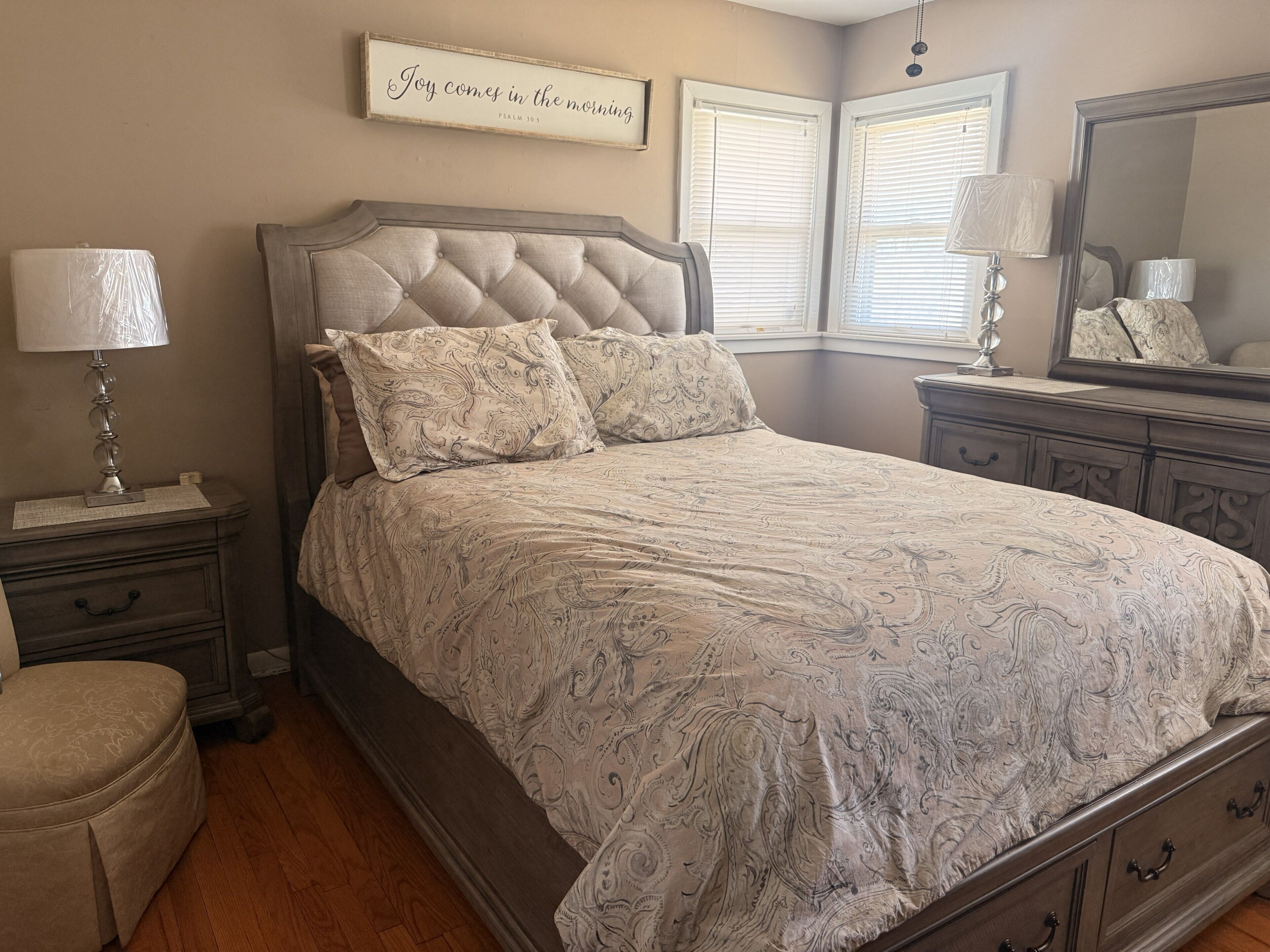 A bedroom with a tufted headboard, a patterned bedspread, a bedside table with a lamp, a nightstand, a dresser, and a mirror.