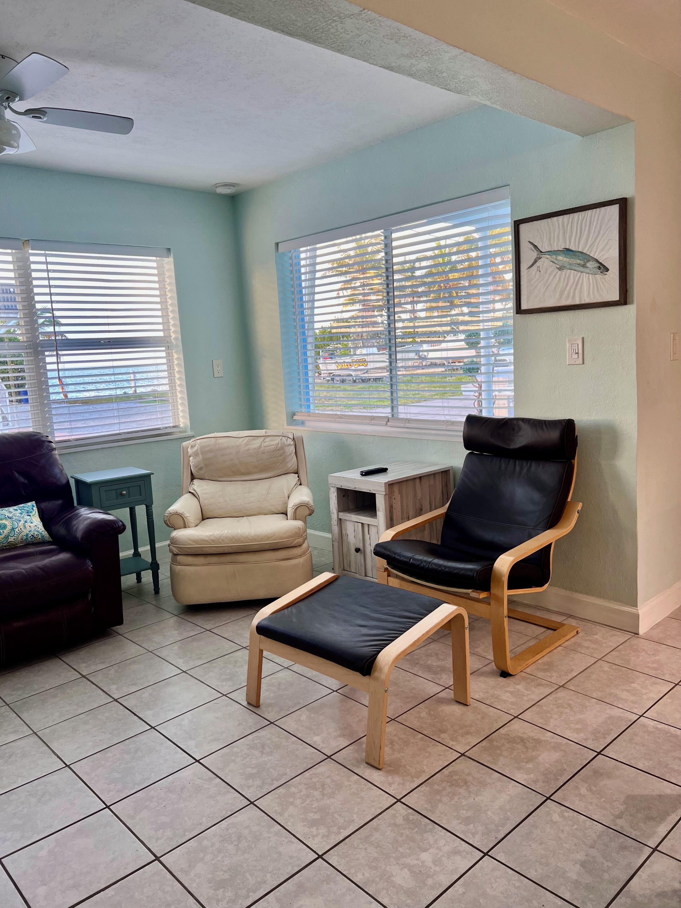 A room with a ceiling fan, two windows with blinds, a framed fish picture, a beige armchair, a black leather chair, a small wooden table, and a dark leather sofa.