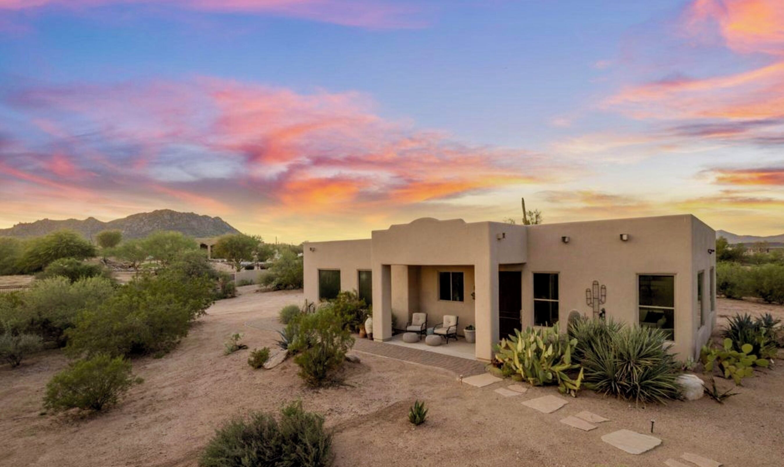 A modern building with a flat roof, a patio area with seating, and a desert landscape with sparse vegetation.