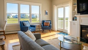 Ein Wohnzimmer mit einer Couch, Sesseln, einem Glastisch, einem Kamin und großen Fenstern mit Blick ins Freie.
