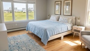 Ein Schlafzimmer mit Bett, Nachttisch, Fenster und Blick auf Häuser und Grünflächen.