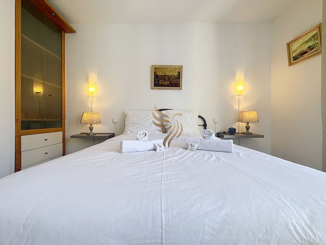 A hotel room with a large bed, two bedside tables with lamps, a wardrobe, and a painting on the wall.