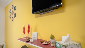 Ett rum med en gul vägg, en TV som hänger på väggen, ett rött bord med en tallrik mat, en näsdukslåda, en liten växt och en skylt med information om Wi-Fi.