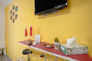 A room with a yellow wall, a mounted TV, a red table with a plate of food, a tissue box, a small plant, and a sign with WiFi details. - Apartment 'Kaktus 2 - Music' with Wi-Fi and Air Conditioning (Bari)