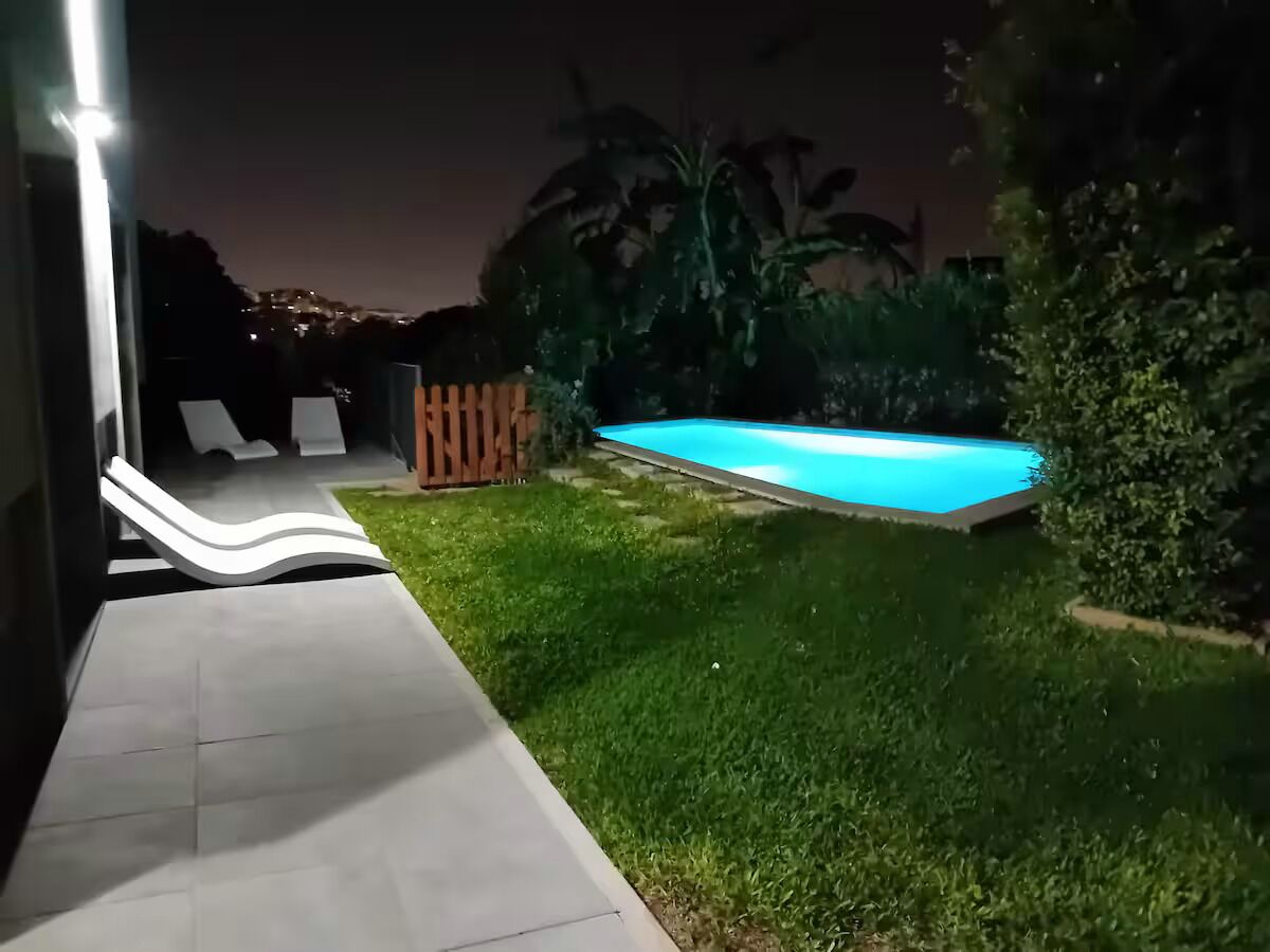 Vista nocturna de un área de piscina con deck de baldosas, cerca de madera y abundante vegetación.