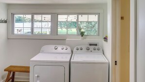 A laundry room with two white front-loading washers and dryers, a wooden bench, a window with a view of a yard, and a door to the right. - Little Farm House with Shaded Front Porch, Fire Pit, Covered Parking (Cross Plains)