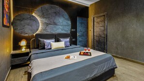 Quarto de hotel moderno com uma cama grande, cabeceira com design circular, mesa de cabeceira com luminária e bandeja com frutas e copos.