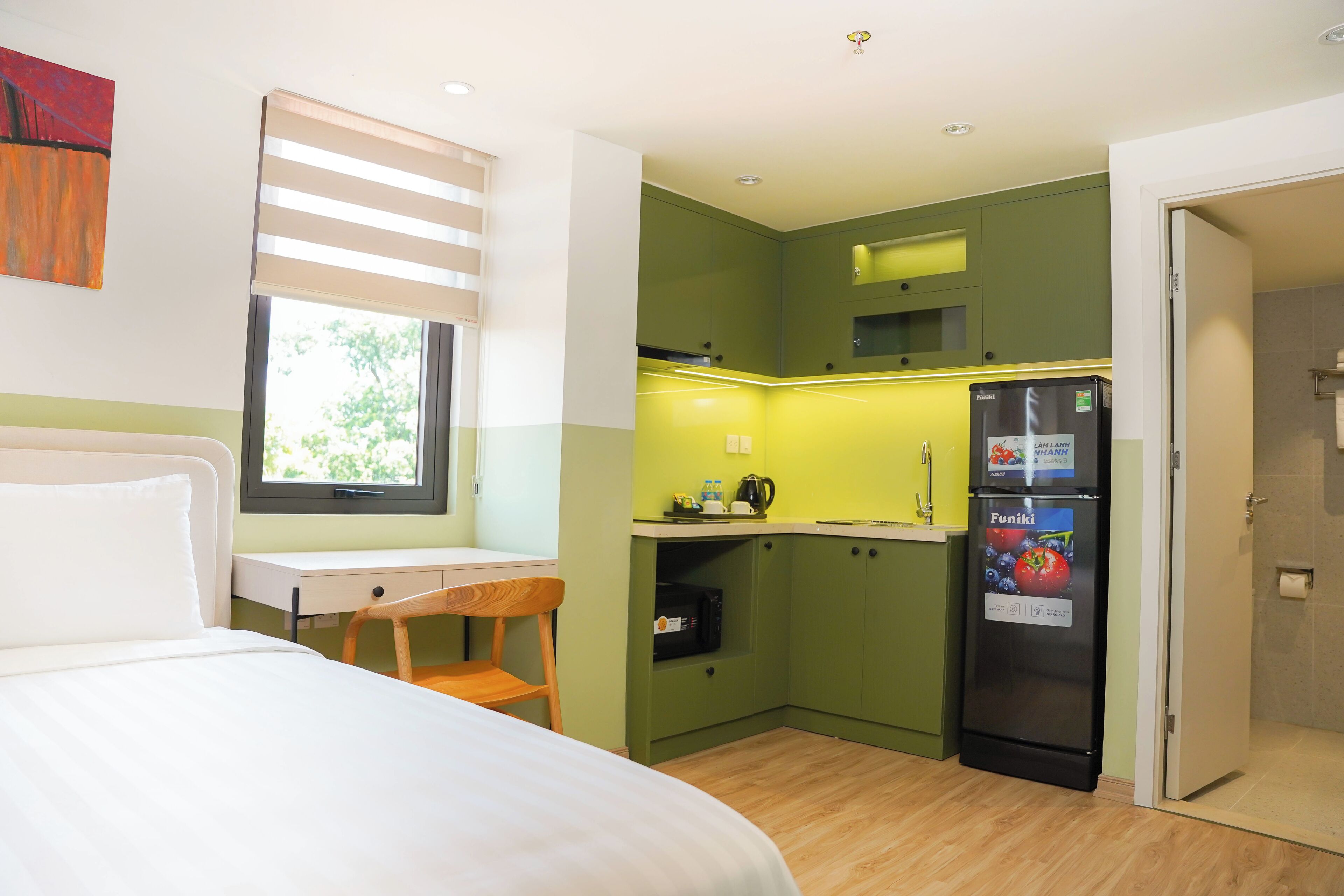 A compact room with a bed, a small desk, a chair, a kitchenette with a refrigerator, and a window.