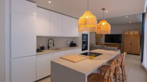 Una cocina moderna con armarios blancos, una isla central, sillas de madera y lámparas colgantes.
