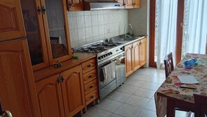 Une cuisine avec des meubles en bois, une cuisinière à gaz, un évier et une table à manger recouverte d’une nappe à motifs floraux.