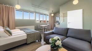 Ein moderner, hell eingerichteter Raum mit Bett, Sofa und Essbereich, ausgestattet mit großen Fenstern und Blick auf die Stadt.