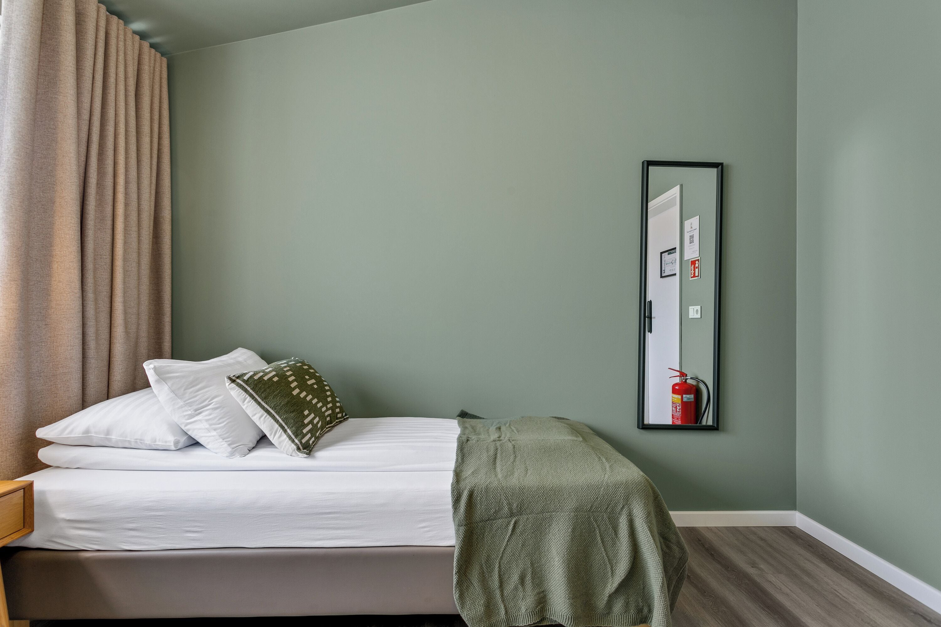 A neatly made bed with white linens and a green throw blanket, a wooden nightstand, a beige curtain, and a wall-mounted mirror reflecting a door and a fire extinguisher.