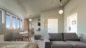 Ein modernes Zimmer mit einem Bett, einem grauen Sofa, einem Esstisch mit Stühlen, einer kleinen Küchenzeile und großen Fenstern.