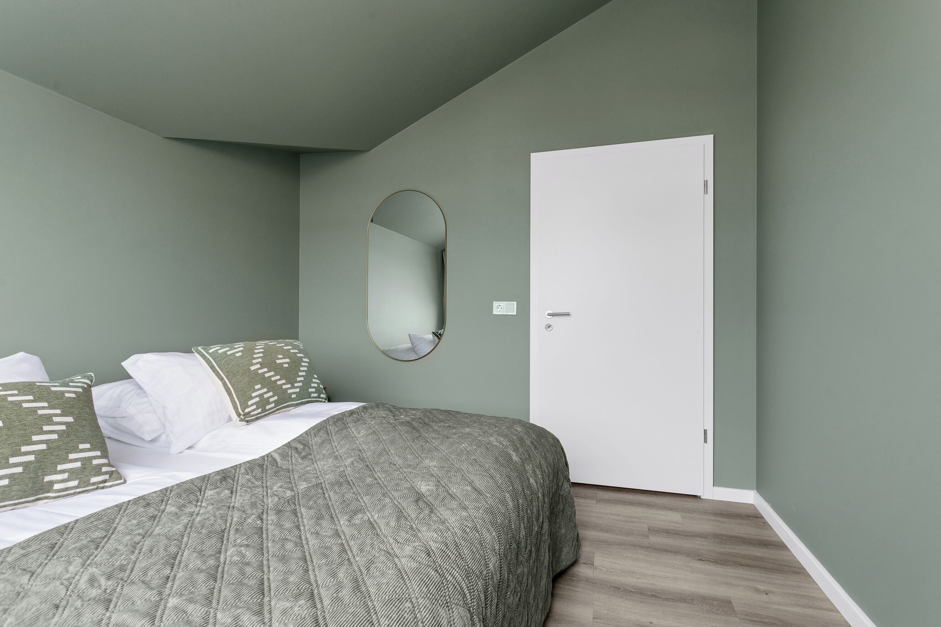 A bedroom with a bed, pillows, a mirror, a door, and a light switch.