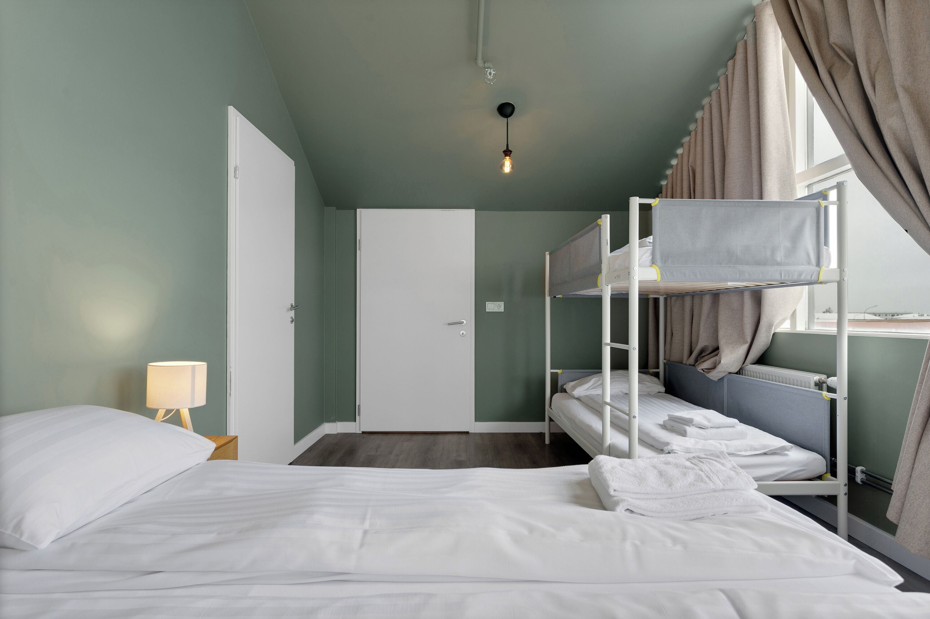 A room with bunk beds, a single bed, a door, a lamp, and a window with curtains.