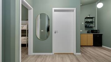 A room with a bed, a door, a mirror, a kitchen area with a microwave, and a shelf with kitchenware.