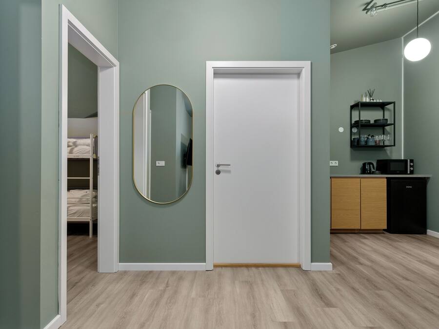 A room with a bed, a door, a mirror, a kitchen area with a microwave, and a shelf with kitchenware.