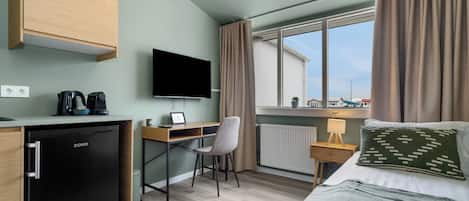 A compact hotel room with a kitchenette, a bed, a desk with a chair, a TV, and a window with curtains.