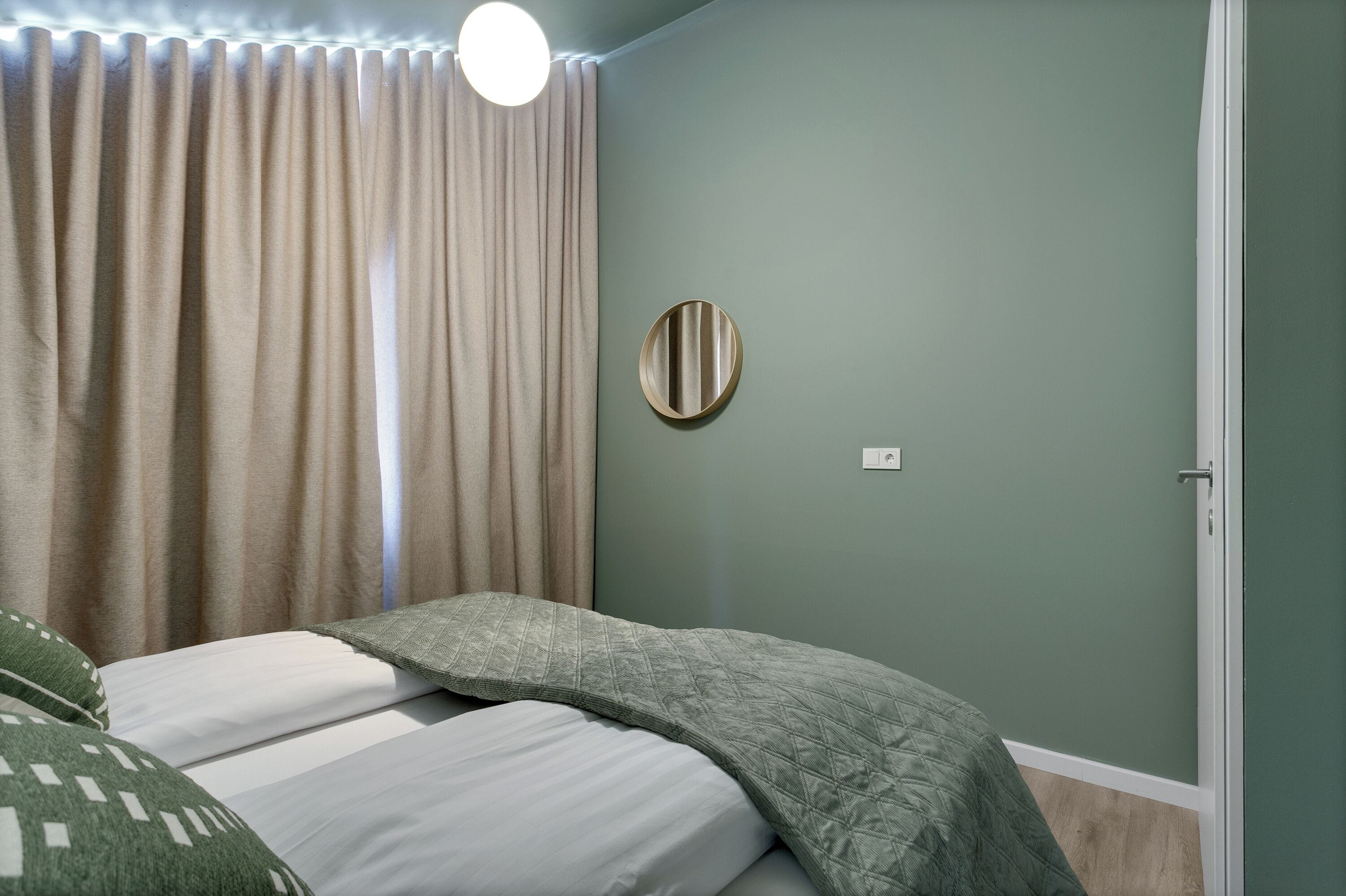 A bedroom with a bed, curtains, a round mirror, and a door.