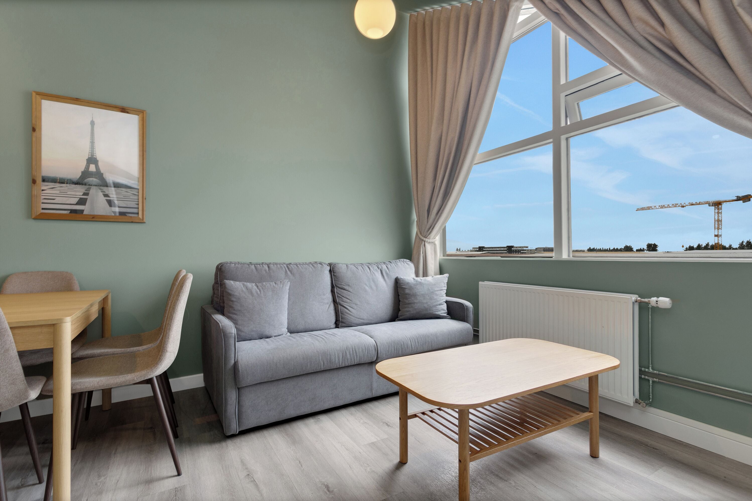 Una habitación con un sofá gris, una mesa de madera, una silla y una ventana con cortinas.