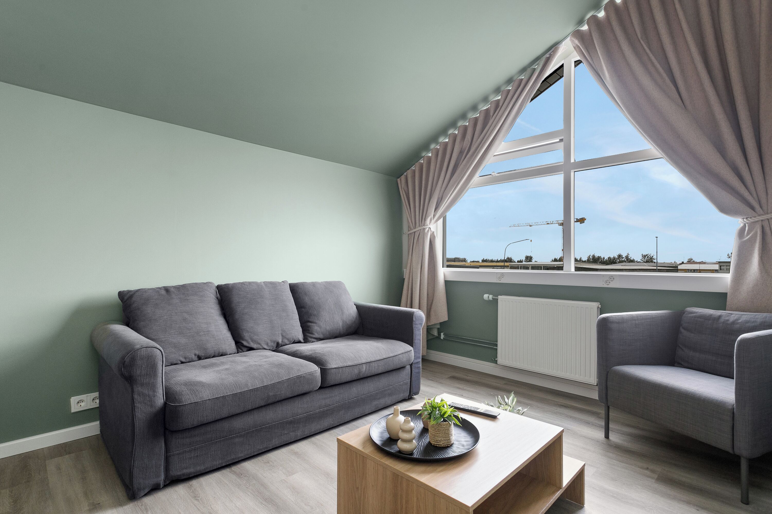 A modern living room with a grey sofa, a wooden coffee table, and a view of a construction site through a large window.