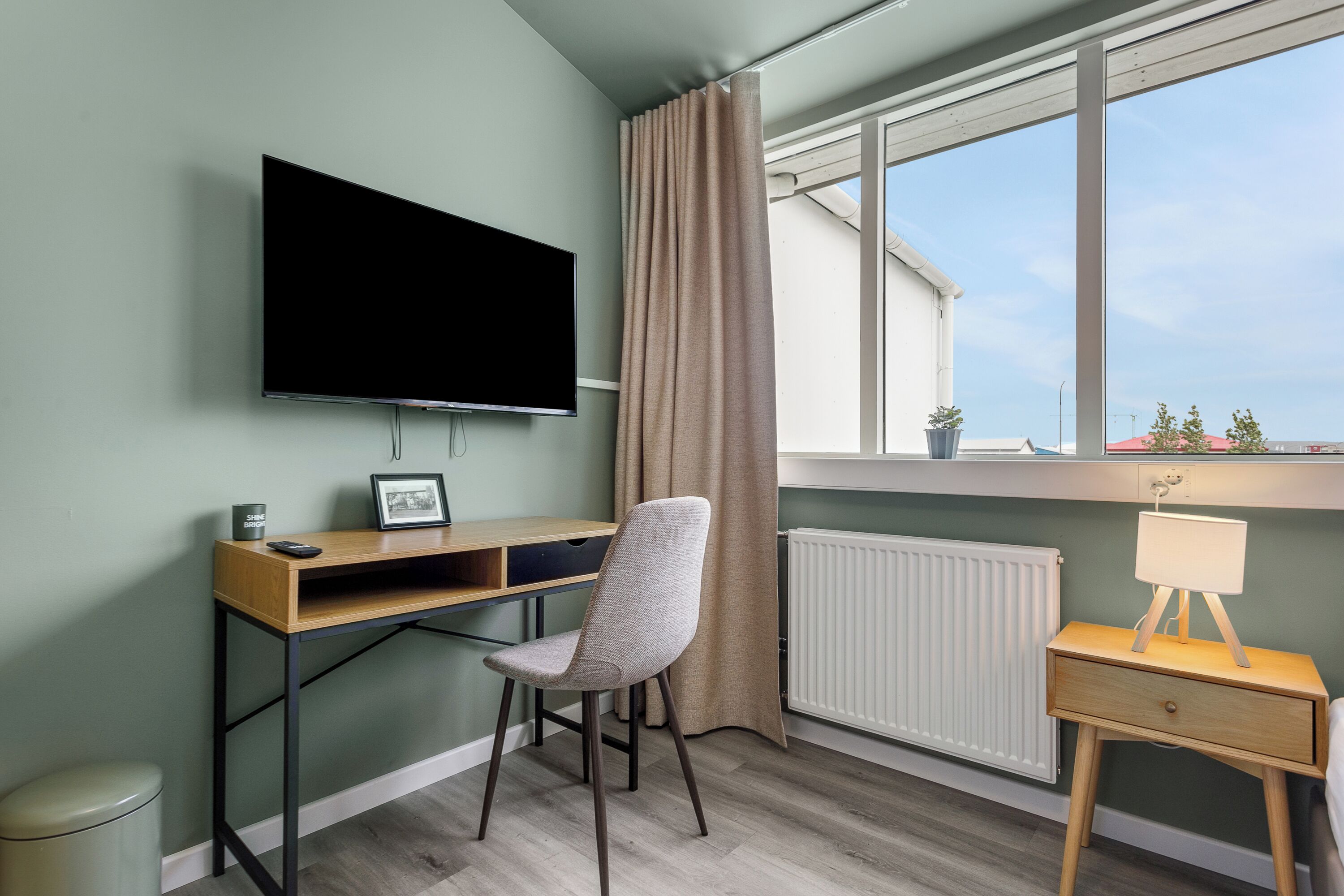 Une pièce comprenant un bureau, une chaise, un téléviseur, un radiateur, une fenêtre avec des rideaux et une petite table avec une lampe.