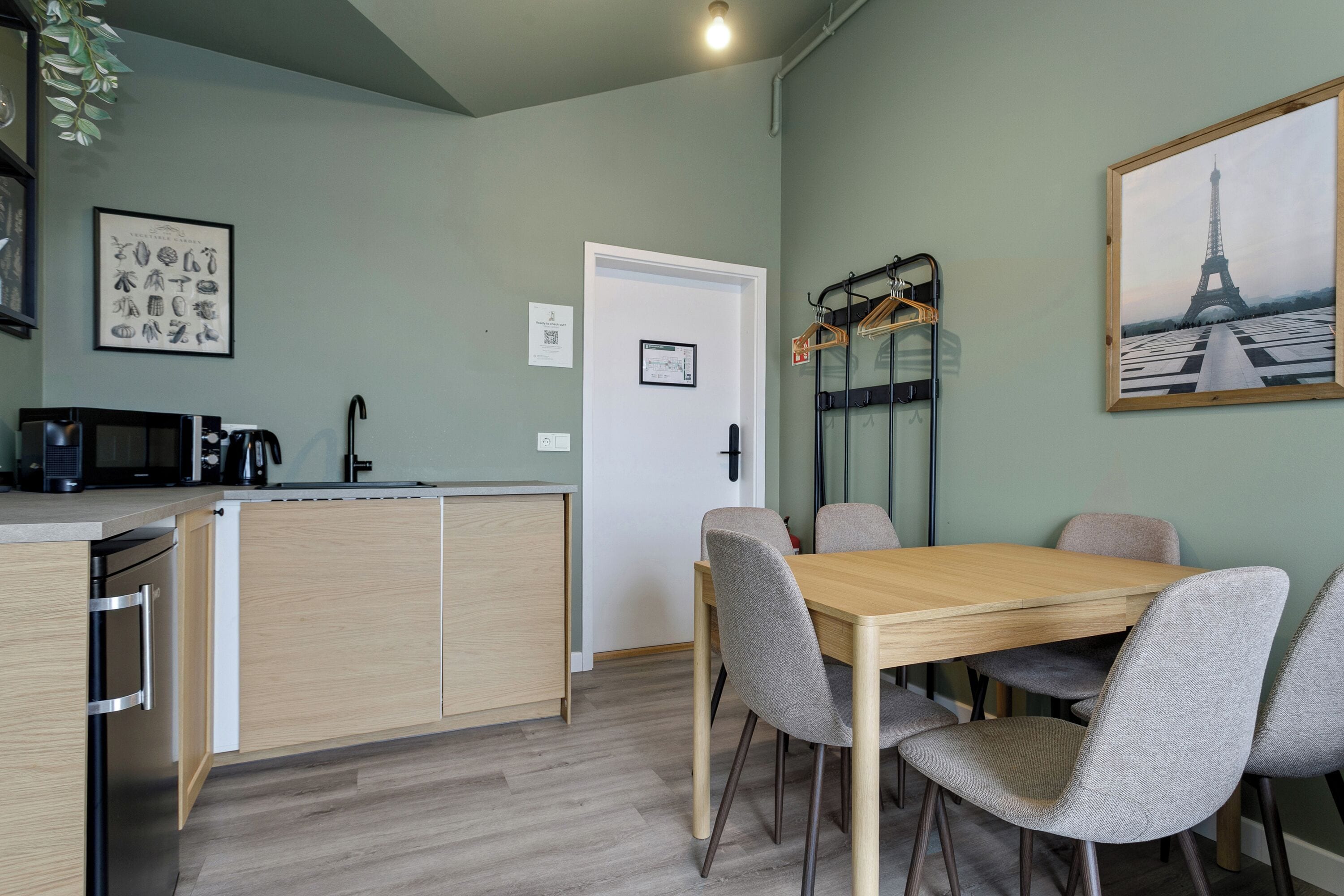 A modern kitchen with a built-in microwave, a kettle, and a coffee maker. There is a dining area with a wooden table and four chairs. A framed picture of the Eiffel Tower is on the wall.