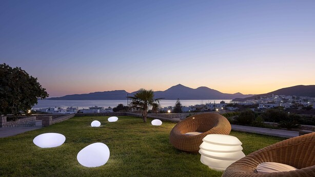 Un espacio exterior con césped, asientos y vista a una ciudad costera al atardecer.