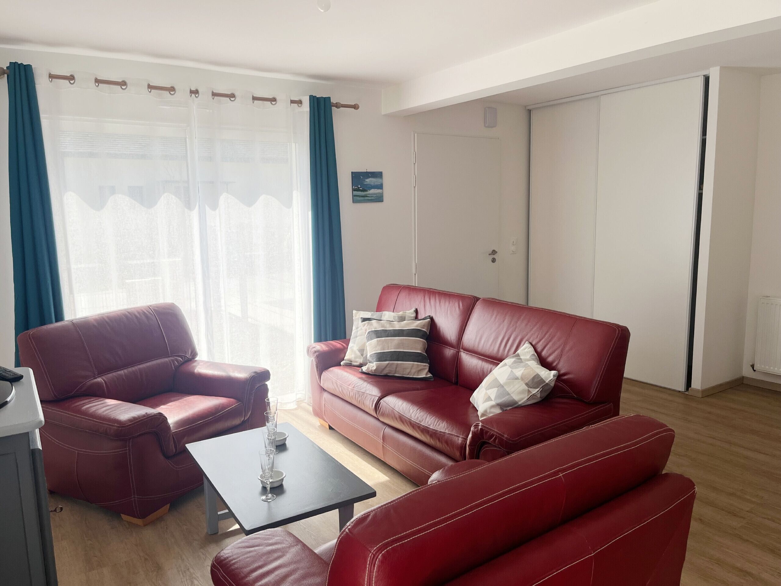 Un salón con un sofá de cuero rojo, una mesita con vasos y una ventana con cortinas.