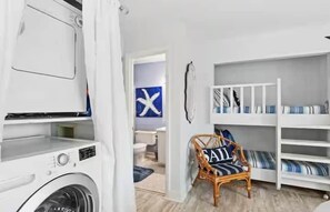 A laundry room with a washing machine, a wicker chair, and a bunk bed with striped bedding. - Beachfront with Pool No Elevators, Just The BEACH (Panama City Beach)