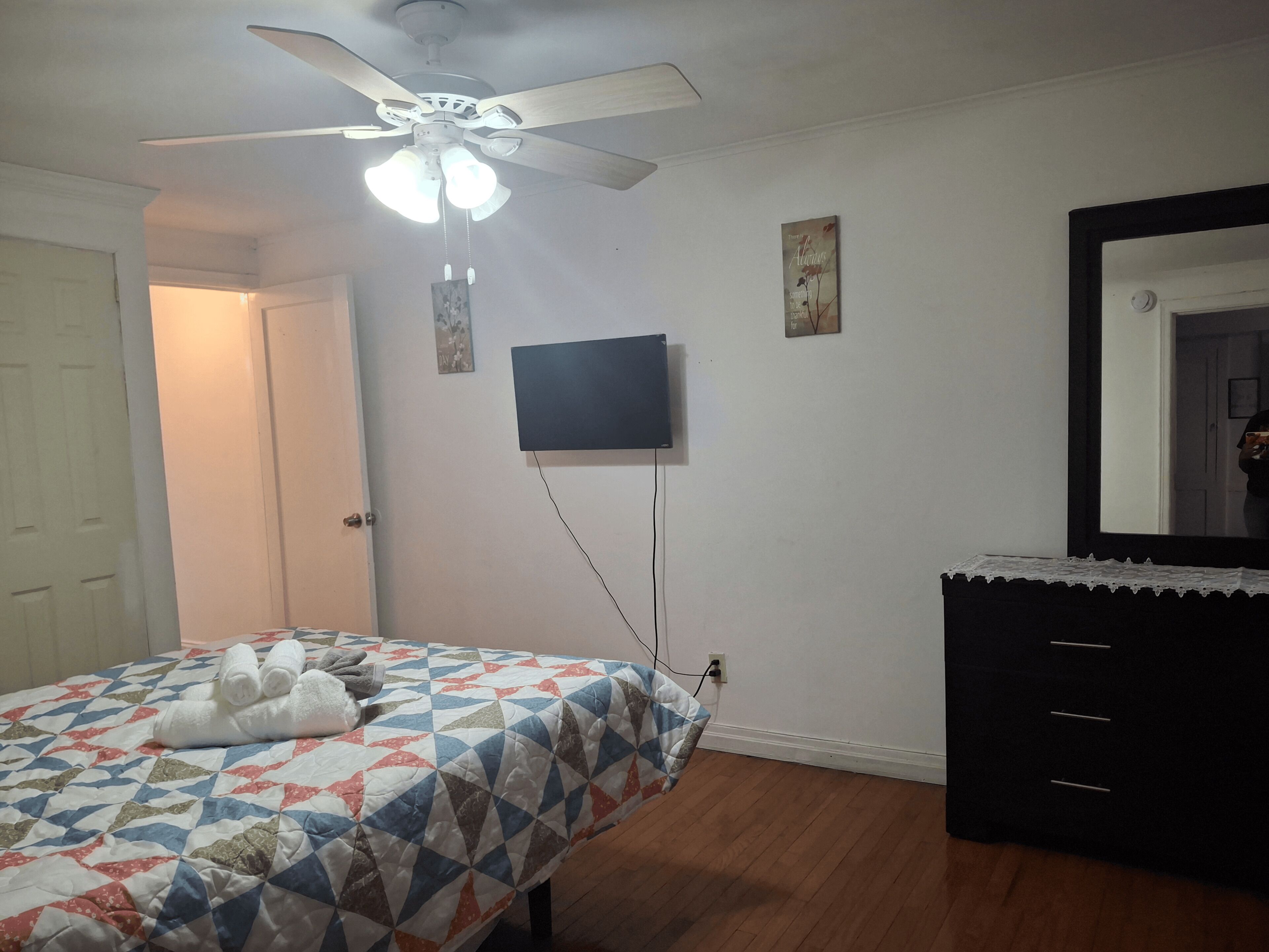 Quarto com cama, ventoinha no teto, televisão na parede, cómoda e porta.