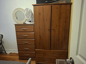 A wooden wardrobe with a mirror and a fan on top, a small desk with a lamp, and a door with a handle.