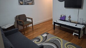 A living room with a sofa, a chair, a painting of a cityscape, a television, and a small table with a placemat. - Tranquil 1-bedroom apartment share with AC in vibrant Brooklyn Near B8 Bus Stop (Brooklyn)