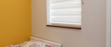 A bedroom with a bed, a window with blinds, and a yellow wall.
