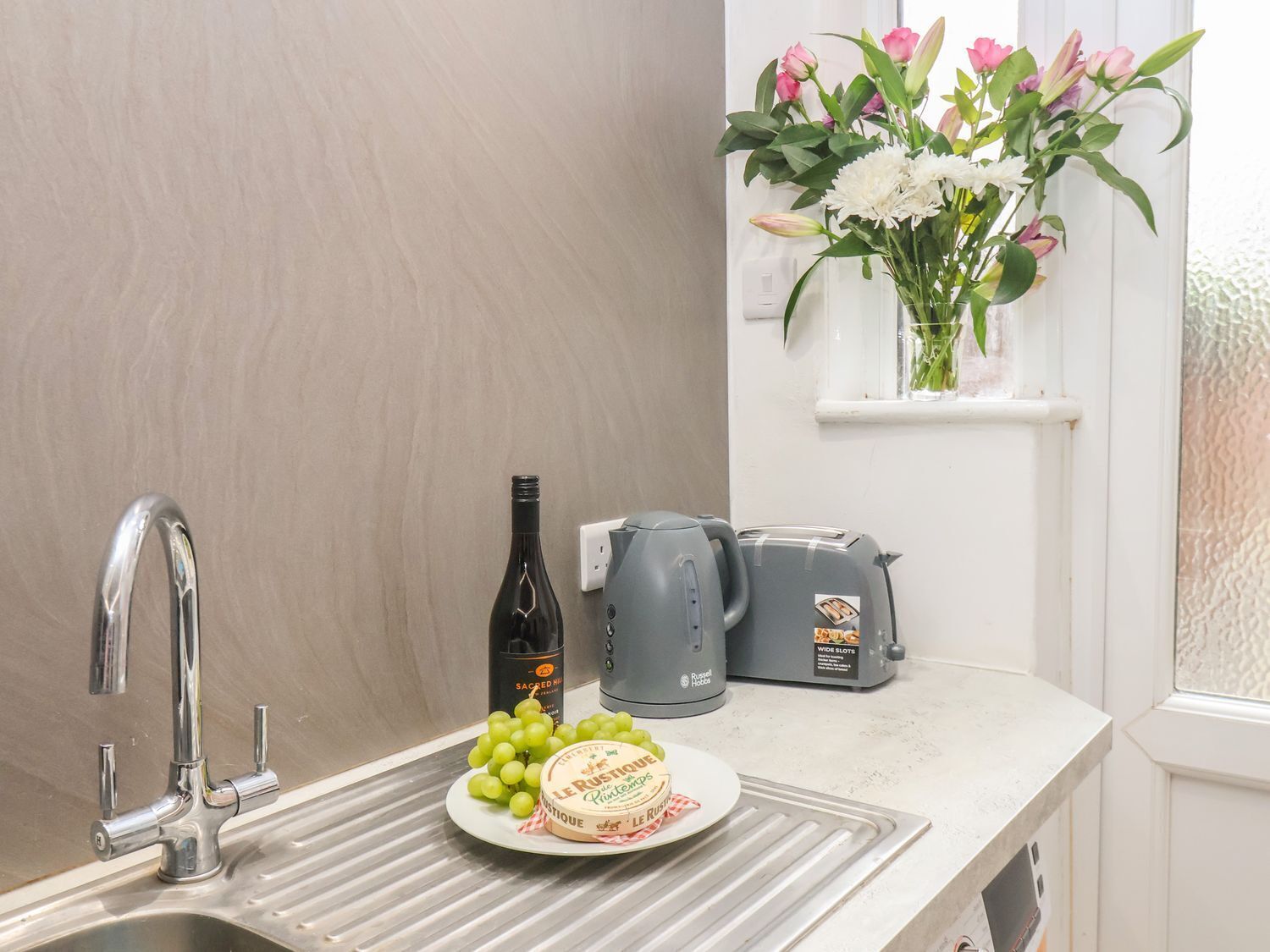 A kitchen with a stainless steel sink, a wine bottle, a plate of grapes, a cheese plate, a kettle, and a toaster.