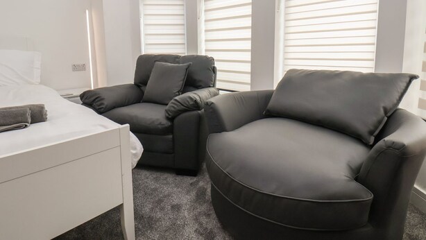 A modern hotel room with a bed, two dark grey sofas, and large windows with blinds. - Frankies Seaview 4 (Bridlington)