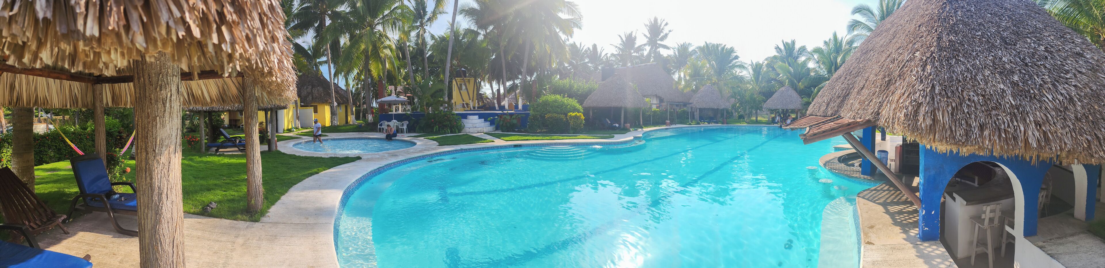 Una piscina con estructura de techo de paja, rodeada de palmeras y un césped bien cuidado.