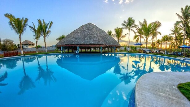 A swimming pool with a thatched-roof structure, palm trees, and a clear sky. - Hotel Cafe Del Sol (Monterrico)