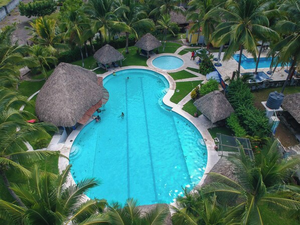 A large oval-shaped swimming pool surrounded by palm trees and thatched-roof structures. - Hotel Cafe Del Sol (Monterrico)