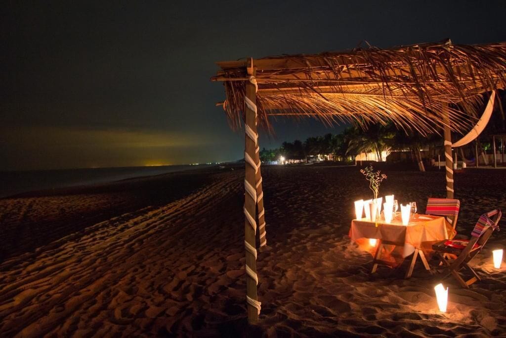 A beach setting with a thatched structure, a table with candles, and beach chairs.