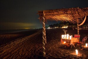 A beach setting with a thatched structure, a table with candles, and beach chairs. - Hotel Cafe Del Sol (Monterrico)