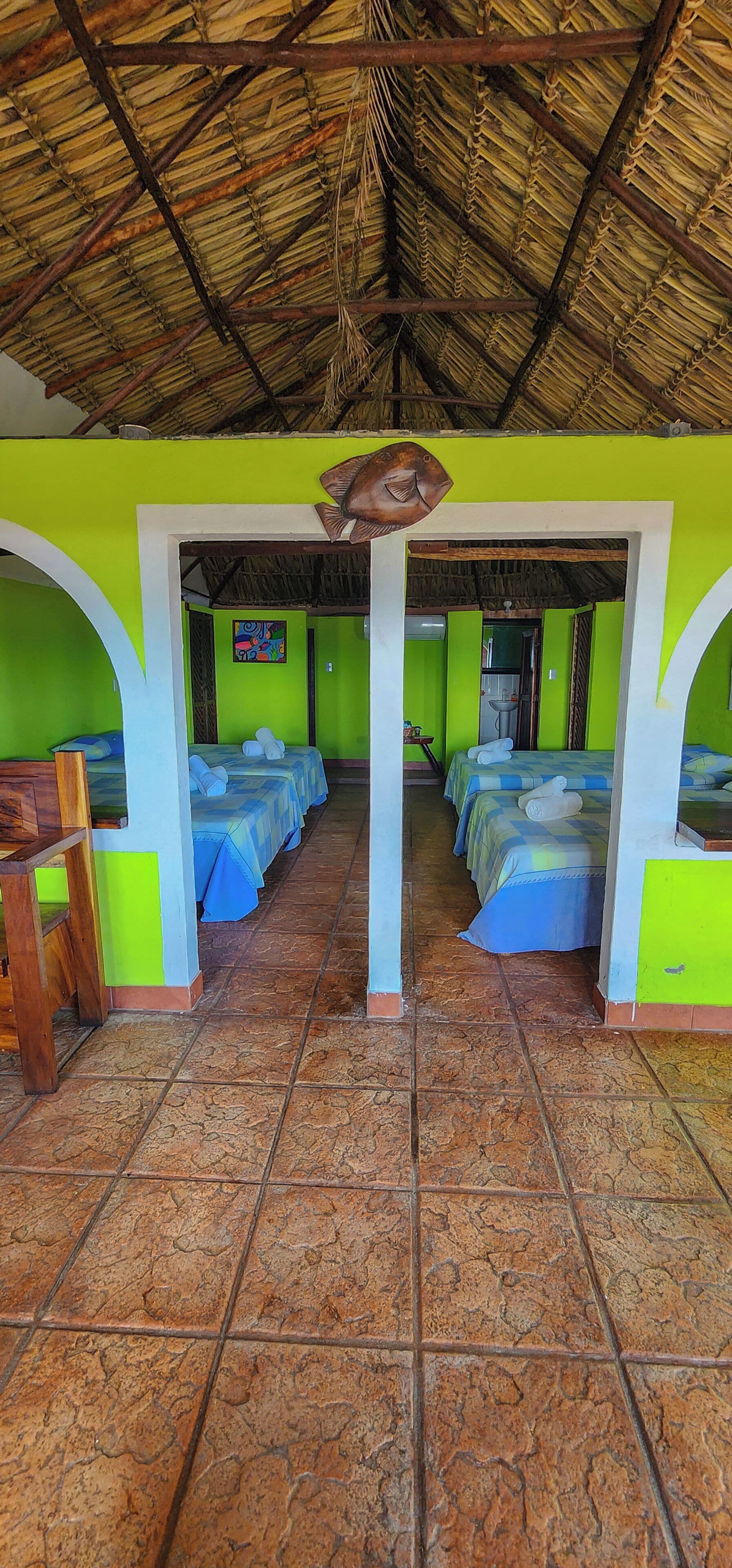 A room with a thatched roof, green walls, and tiled flooring. There are two beds with blue bedding, a wooden chair, and a fish decoration on the wall.