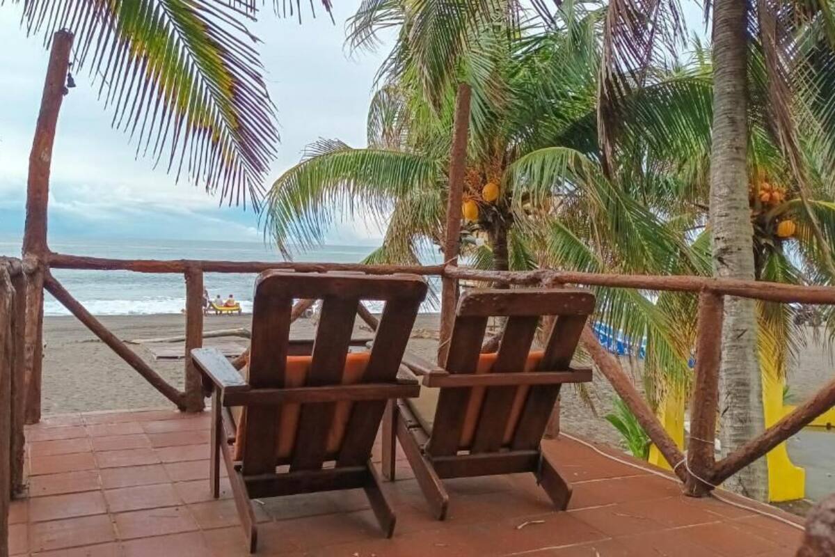Una terraza de madera con dos sillas que da a una playa, palmeras y vistas al océano.
