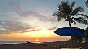 A beach scene with a sunset, a boat labeled 'Cafe del Sol,' and a wooden deck with a blue umbrella. - Hotel Cafe Del Sol (Monterrico)