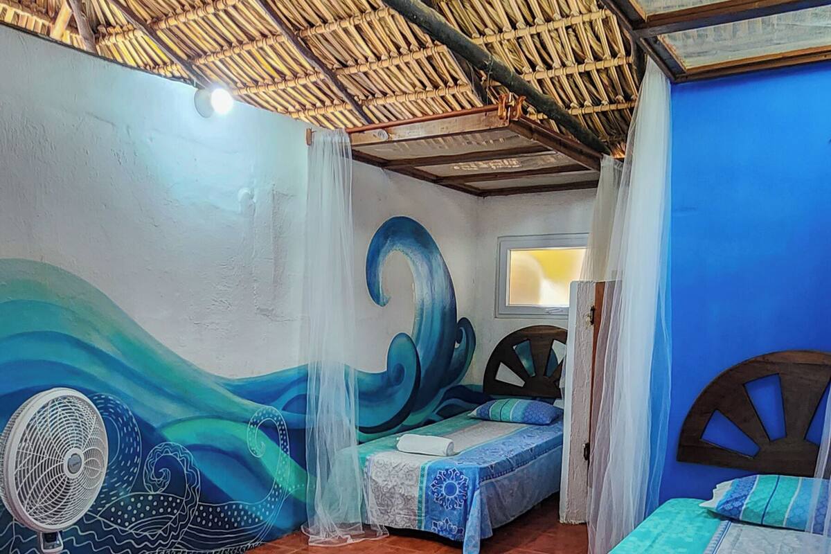 Una habitación con piso de baldosas, dos camas con ropa de cama azul y blanca, un ventilador y un mural en la pared.