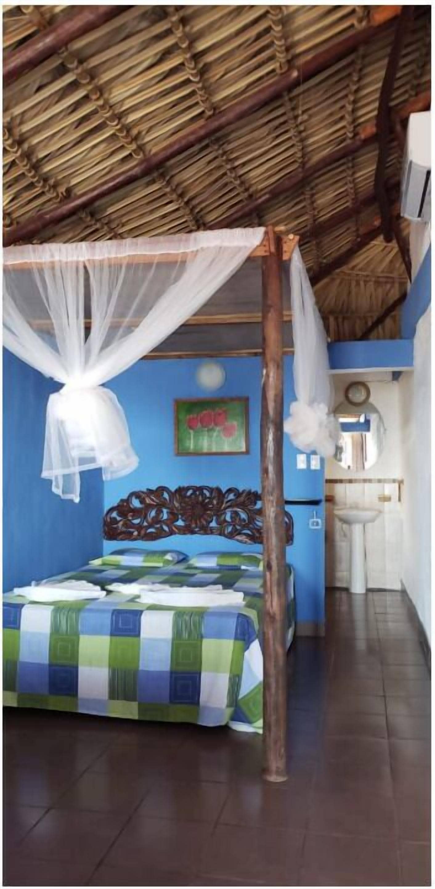 Una habitación con una cama bajo una mosquitera, cabecera de madera, pared azul, un cuadro, un lavamanos y un reloj.