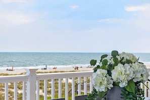 Ein Balkon mit Blick auf Strand und Meer, einem weißen Geländer und einer Vase mit weißen Blumen.