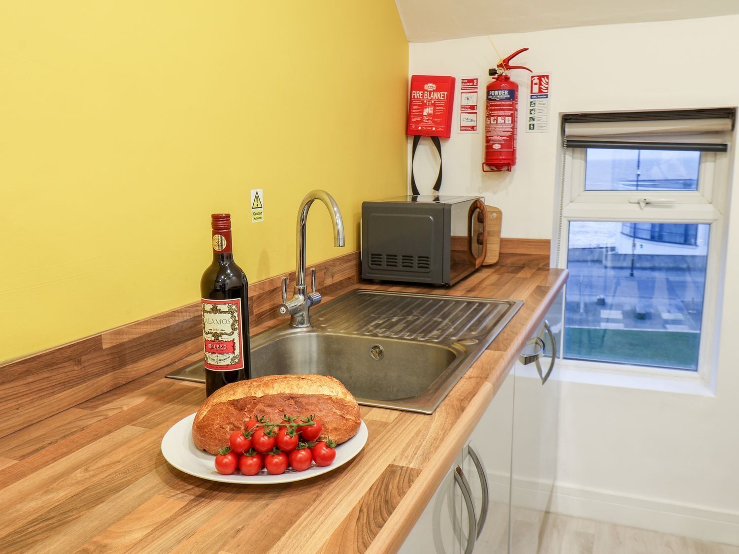 A kitchen with a bread loaf, tomatoes, a bottle, a sink, and fire safety equipment.